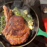 Bone-in Ribeye Steak searing in a cast iron skillet with garlic and herbs.